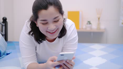 Mujer-Asiática-Mirando-Y-Tocando-El-Teléfono-Inteligente-Relájese,-Disfrute-Y-Sonría-Con-Las-Redes-Sociales-En-Línea-En-El-Dormitorio