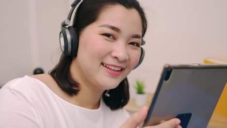 Schöne-Asiatische-Frau-Schaut-Kamera-Und-Lächelt,-Entspannt-Sich-Und-Hört-Musik-Per-Tablet-Mit-Kopfhörern-Auf-Dem-Bett-Zu-Hause-Lifestyle--Und-Vergnügungskonzept