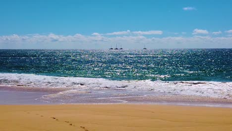 HD-Hawaii-Kauai-backlit-slow-motion-static-wide-shot-of-sparkling-ocean-waves-washing-up-on-beach-with-second-boat-of-four-boats-passing-first-boat-near-center-of-frame-with-partly-cloudy-and-blue-sky
