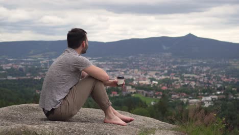 Caucasian-man-sitting-on-rock-and-drinking-coffee-from-paper-cup,-beautiful-view-in-background