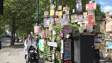 A-man-with-a-child-in-pushchair-passes-a-huge-memorial-wall-of-cardboard-signs-showing-support-of-healthcare-staff-during-the-Coronavirus-outbreak-line-the-edge-of-a-park-in-the-East-End-of-London
