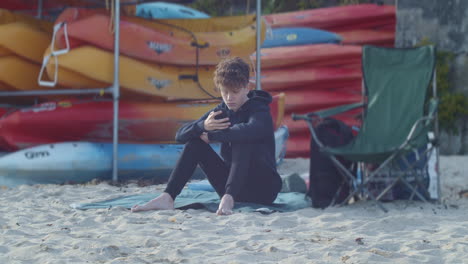 Teen-boy-sits-on-beach-with-phone-near-kayak-rentals