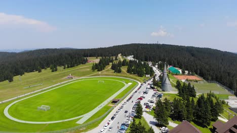 Hotel-Rogla-resort-soccer-field-with-Podruznicna-Church-to-the-right-during-spring-season,-Aerial-flyover-shot