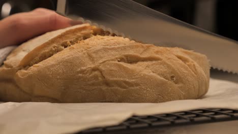 Person-cutting-through-freshly-baked-loaf-of-sour-dough-bread-with-sharp-serrated-kitchen-knife-on-bench-top