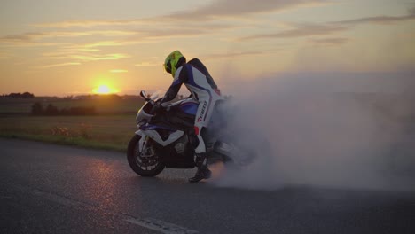 Medium-wide-slow-motion-shot-of-a-motorbiker-wearing-full-body-suit-and-helmet-performing-a-burnout-with-a-white-superbike-during-sunset