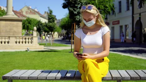 COVID-19-young-beautiful-blonde-teen-women-sitting-on-bench-waiting-and-checking-phone-in-public-city-plaza