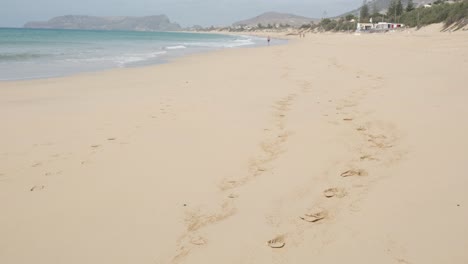Footprints-on-sandy-beach-of-Porto-Santo-island-by-turquoise-ocean-sea-water-waves-rolling-on-shoreline-coast-with-mountain-range-in-background,-Portugal,-handheld-pan-up
