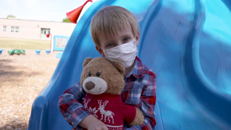 Little-boy-in-a-mask-cuddling-his-teddy-bear-at-a-playground