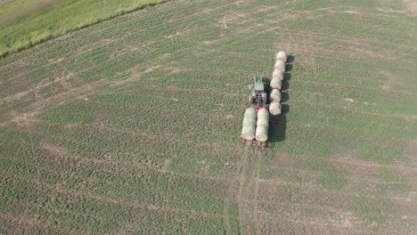 Tractor-with-trailer-picking-up-round-hay-bales-on-green-farmland,-follow-aerial