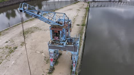 Pan-around-drone-view-of-rusty-crane-covered-with-graffiti-and-located-on-concrete-pier-near-water-in-city