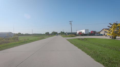 POV-driving-on-a-rural-highway-thru-the-edge-of-a-small-tow-past-a-lot-with-large-farm-equipment---Hill,-Iowa