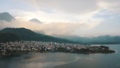 Vista-Aérea-De-Drones-De-Una-Ciudad-Y-Un-Hermoso-Arco-Iris-En-El-Lago-Atitlán,-Guatemala---Paisaje-De-Montañas-Y-Volcanes