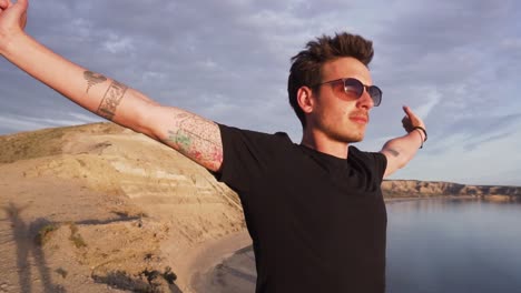 Young-Man-In-Black-Shirt-Wearing-Sunglasses-Standing-With-Open-Arms-On-A-Mountain-Facing-The-Sea-At-Sunset---Medium-Shot