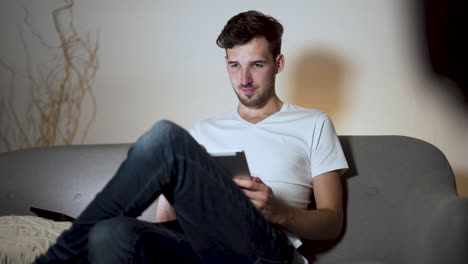 A-young-man-with-a-stubble-and-a-modern-hairstyle,-wearing-a-white-t-shirt-and-jeans,-sitting-bored-at-home-on-a-sofa,-legs-crossed,-smiling-while-browsing-his-tablet,-static-4k