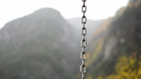 a-chain-from-a-swing-chair-dancing-in-the-rain-during-a-cold-autumn-day,-the-blurry-mountains-behind-offer-a-beautiful-scene