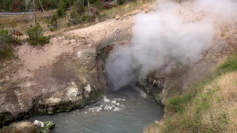 Steam-rises-from-Dragon's-Mouth-Spring-in-Yellowstone-National-Park,-and-a-wave-of-water-is-ejected-from-the-opening