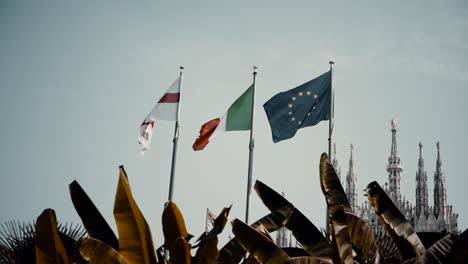 Bandera-De-Inglaterra,-Italiana-Y-Europea-Ondeando-En-El-Viento-Con-El-Famoso-Duomo-Di-Milano-En-Milán,-Italia
