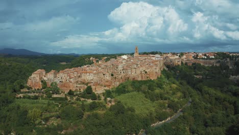Drone-footage-of-the-historic-medieval-town-Pitigliano,-a-masterpiece-of-ancient-architecture-on-a-rock-in-the-idyllic-landscape-of-Tuscany,-Italy-with-green-trees,-blue-hills-and-thunder-storm-clouds