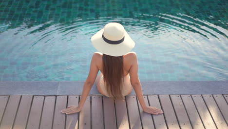 A-young-woman-wearing-a-sun-hat-and-bathing-suit-sits-with-her-back-to-the-camera-on-the-edge-of-a-pool-deck-leans-back-on-her-arms