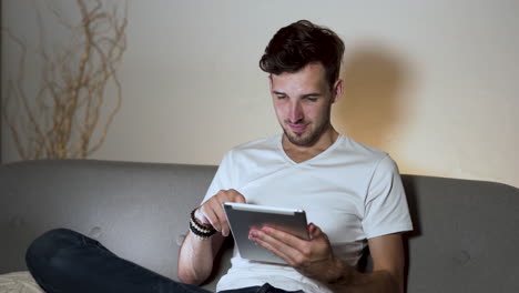 A-young-man-with-a-stubble-and-a-modern-hairstyle,-wearing-a-white-t-shirt-and-jeans,-sitting-at-home-on-a-sofa,-legs-crossed,-nodding-his-head-in-approval-while-swiping-on-his-tablet,-static-4k