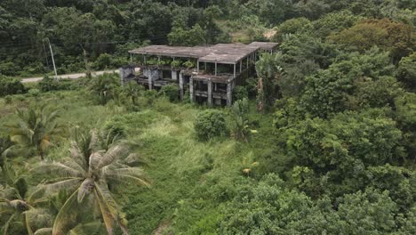 Aerial-backwards-dolly-shot-of-derelict-abandoned-small-hotel-building-on-a-tropical-island-with-palm-trees