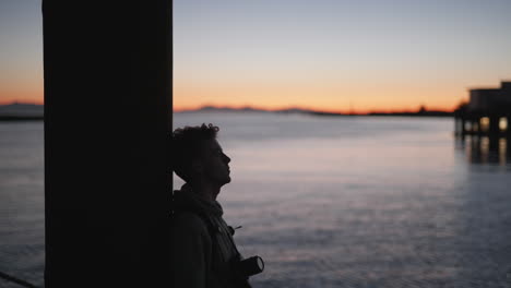 Silhouette-of-young-man-relaxing-with-camera-on-pier,-sunset-over-water