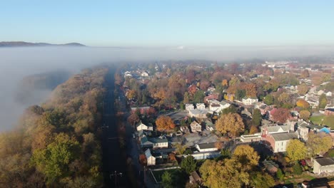Rising-aerial-reveals-town-in-colorful-autumn-fall-foliage,-surrounded-by-fog-and-clouds-in-morning-golden-light