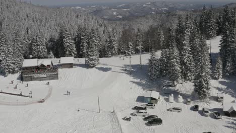 Kope-ski-resort-in-the-Pohorje-mountains-with-visitors-sledding-near-a-cafe-cabin,-Aerial-tilt-down-approach