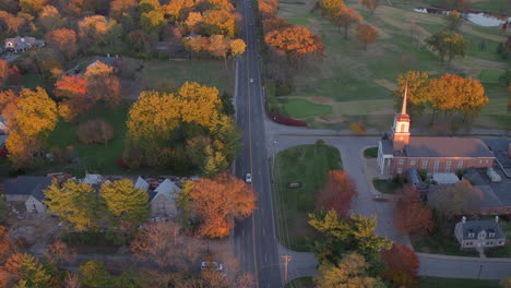Aerial-of-beautiful-Autumn-landscape-in-Ladue-of-a-road-with-cars-alongside-trees-at-peak-Fall-color-and-a-little-church-in-view-at-golden-hour