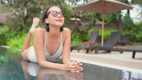Lying-on-her-stomach-and-kicking-her-feet-slowly-in-the-air,-an-attractive-young-woman-in-sunglasses-enjoys-the-amenities-a-resort-has-to-offer