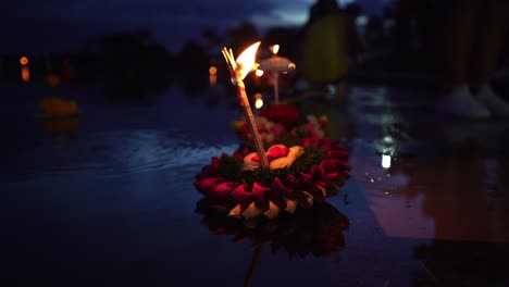Loy-Krathong-Festival:-Colorful-Krathong-leaning-left,-floating-on-the-dark-blue-water-on-the-lake-with-a-burning-candle-and-instance-on-top