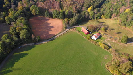 Aerial-view-of-Red-house-surrounded-by-autumn-forest-and-green-field,-Sweden