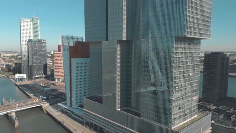 An-aerial-shot-of-a-skyscraper-along-the-shore-of-a-water-channel-and-revealing-the-city-of-Rotterdam,-Netherlands-beyond