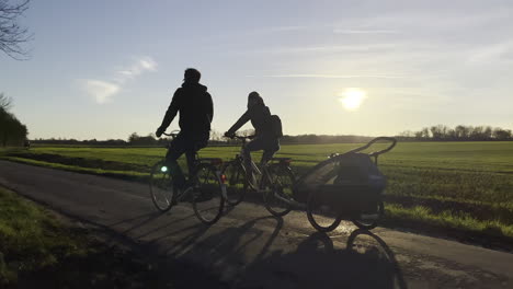 Zeitlupenaufnahme,-Die-Die-Silhouette-Einer-Familie-Auf-Fahrrädern-Mit-Einem-Kindertransporter-Zeigt,-Der-Während-Des-Sonnenuntergangs-Auf-Der-Landstraße-Fährt