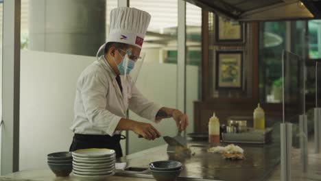 Head-Chef-In-Mask-And-Face-Shield-Mixing-Rice-Dish-At-Griddle-Plate-In-Restaurant-During-Pandemic