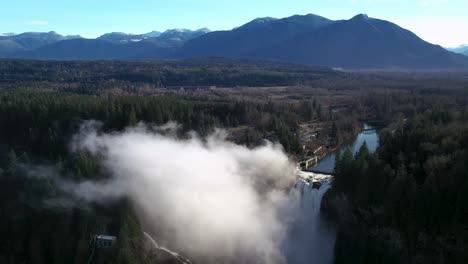 Aerial-orbiting-shot-of-foggy-waterfall-surrounded-by-forest-and-mountain-range-in-background