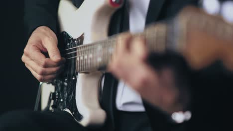 Close-up-of-a-professional-musician-in-a-black-suit-playing-chords-on-a-white-electric-stratocaster-guitar-during-a-live-session-with-a-dark-background