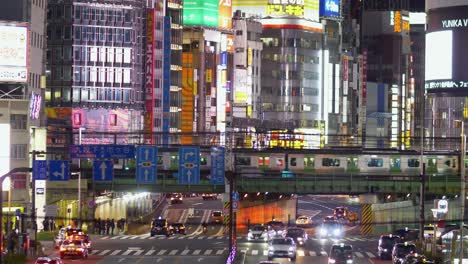 Elevated-night-view-overlooking-traffic,-crossings-and-JR-trains-passing-over-a-bridge,-with-Kabukicho-in-the-background,-in-Shinjuku,-Tokyo