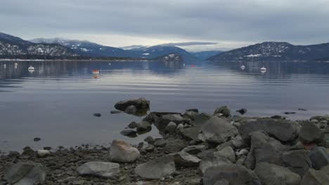 Small-waves-rooling-at-stones-in-Shuswap-lake-with-the-rocky-mountains-on-the-background-at-a-cloudy-day