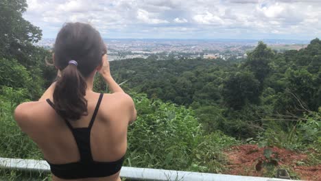 Rear-View-Of-Female-Tourist-Hiker-Taking-A-Panoramic-Picture-Of-The-Cityscape-Using-Her-Smartphone-On-Mountain-Forest-On-A-Sunny-Day-In-Praia-Porto,-Brazil---Medium-Closeup-Shot