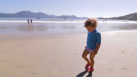 Kamera-Umkreist-Junges-Süßes-Mädchen,-Das-Am-Strand-Mit-Berghintergrund-Und-Ruhigem-Meer-Spazieren-Geht