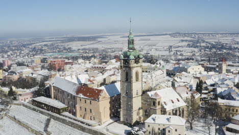 Aerial-view-of-a-town-with-church-by-a-river-bridge-in-falling-snow