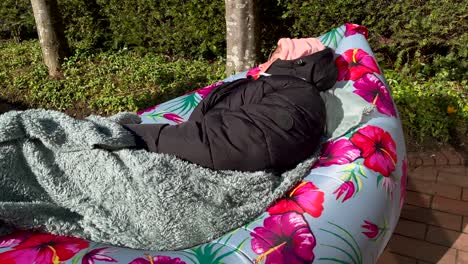 Young-girl-in-jacket-and-blanket-relaxing-in-inflatable-sofa-bed-outdoors-in-garden-during-sunny-day