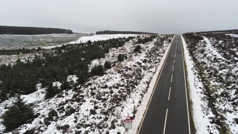 Aerial-following-long-empty-road-into-distance-across-highland-snowy-countryside-moors