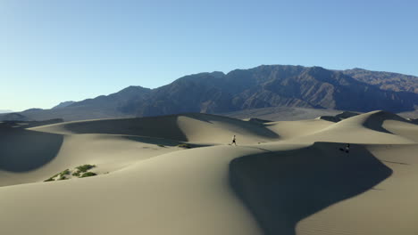 Drone-footage-of-man-walking-through-hot,-dry-sand-dunes-at-sunset,-in-the-desert,-with-mountains-in-background