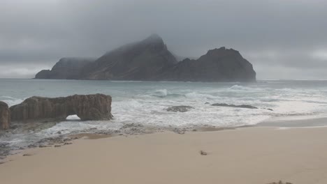 Empty-beach-with-big-rock-shrouded-in-mist-on-background,-Ponta-da-Calheta