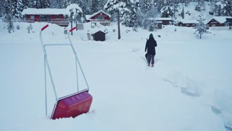 Backview-Of-A-Person-And-Dog-Walking-On-Snow-Covered-Foreground-With-Sled-Shovel-During-Winter-In-Trondheim,-Norway