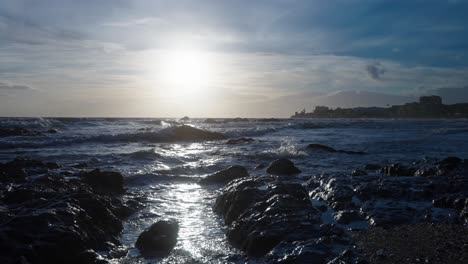 Sun-reflecting-the-waves-that-are-hitting-the-rocks-at-the-beach,-with-the-town-of-Calahonda-on-the-background