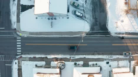 Top-down-aerial-tracking-shot-of-black-car-driving-through-snow-covered-town-in-winter-during-magic-hour-light