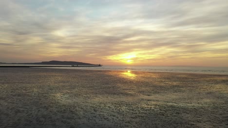 Dublin-Bay,-March-2020,-Drone-gradually-pushes-forwards-towards-South-Bull-Wall-Lighthouse-and-Sunrise,-across-beach-at-low-tide-in-the-early-morning